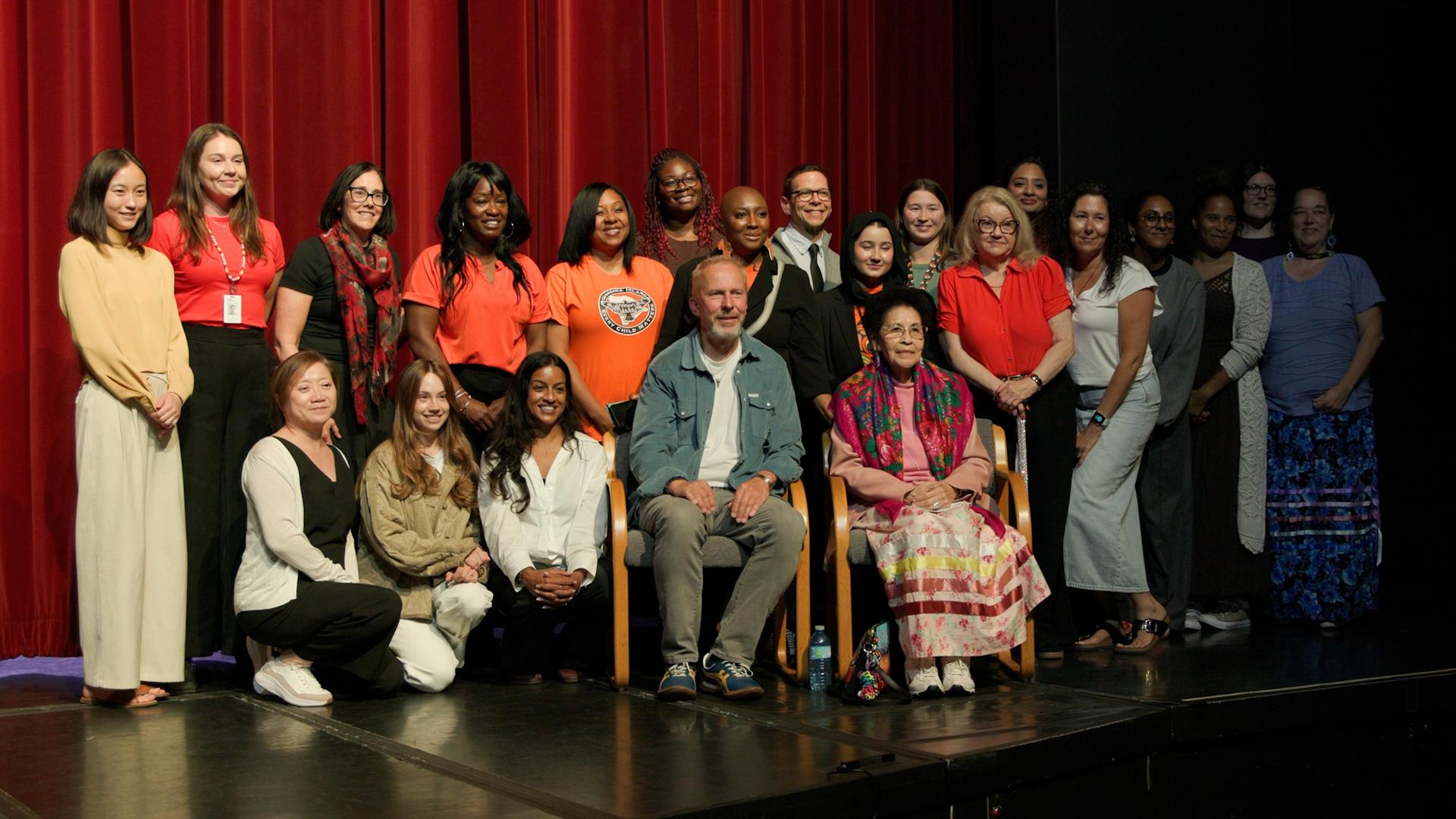 Elder and Director sit on a stage surrounded by staff and students standing or kneeling, some in orange shirts